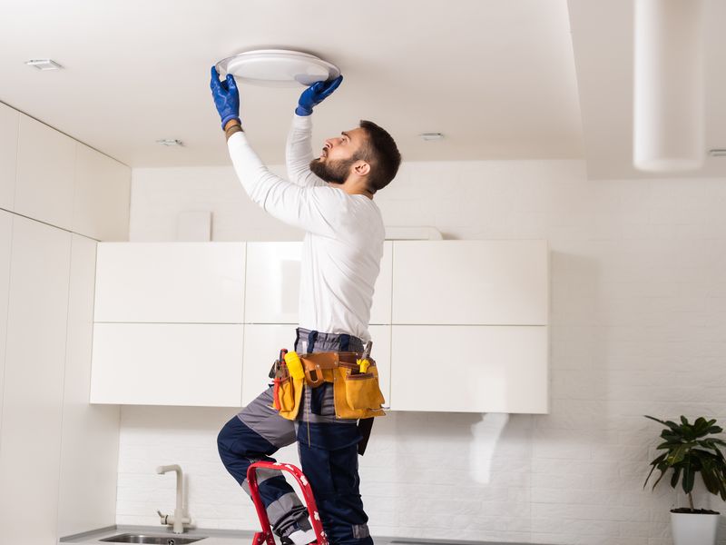 A maintenance worker on a small ladder replaces a ceiling light in a bright kitchen.