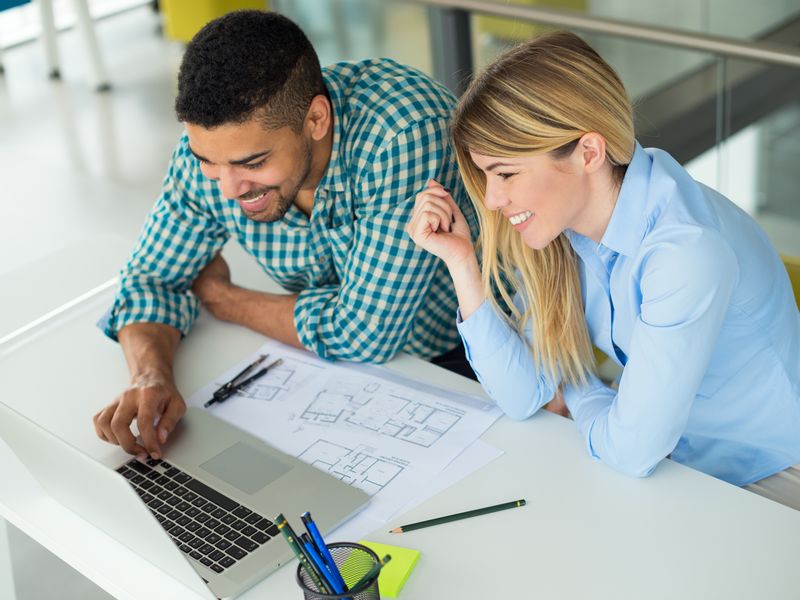 A couple smiling at a laptop with floor plans and drafting tools on the table, planning a remodel.