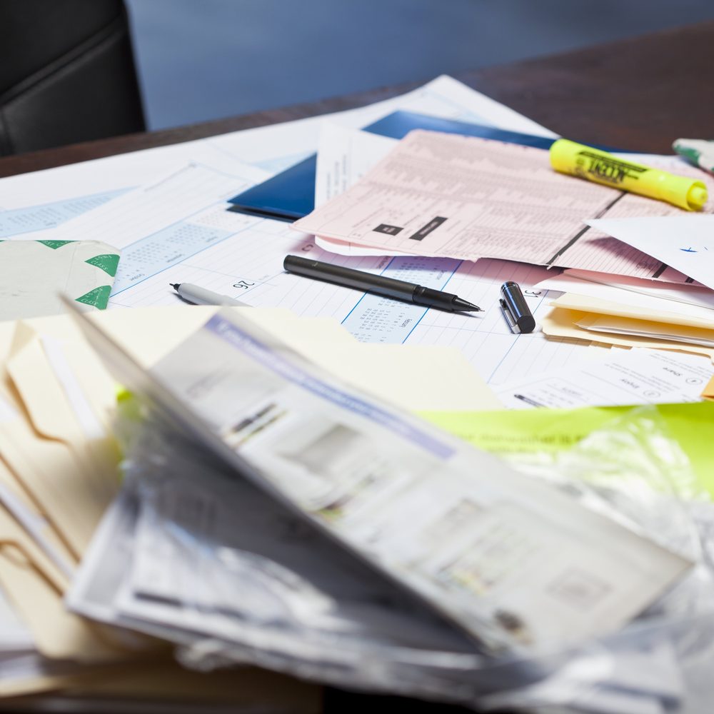A cluttered desk covered in loose papers, envelopes, file folders and a plastic sleeve of documents, showing how records get scattered.